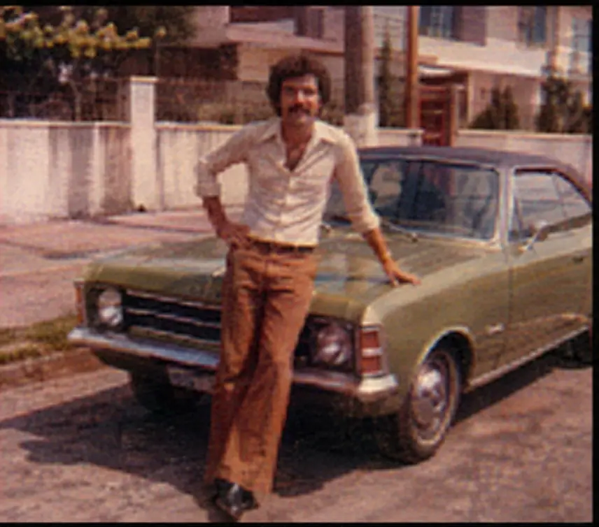 Fotografia analógica antiga de um homem ao lado de um carro, desfocada e em resolução muito baixa